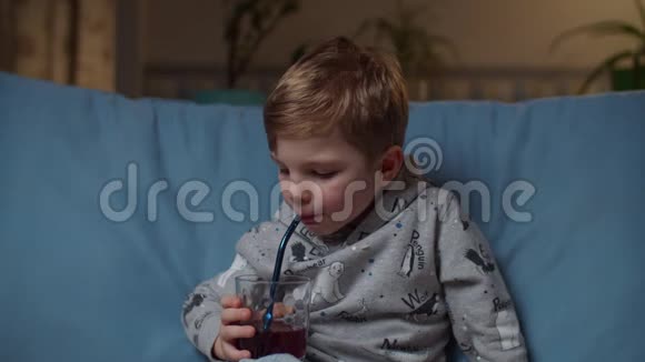 晚上坐在舒适的沙发上喝果汁的学龄前儿童关门小孩喝蓝莓汁而不是视频的预览图