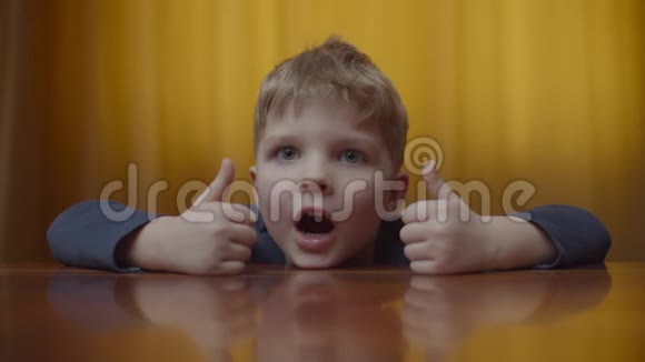 金发学龄前男孩微笑着看镜头的肖像孩子们在家里坐在桌子上微笑的孩子会竖起大拇指视频的预览图