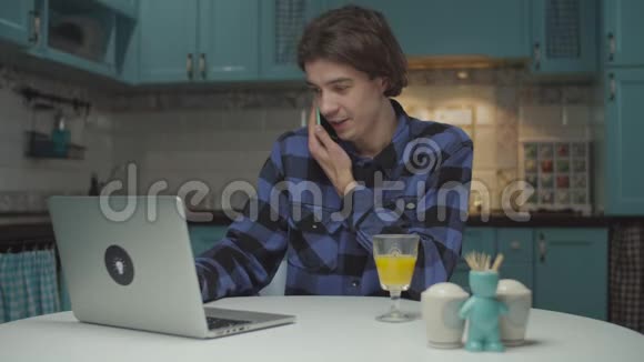年轻的20多岁的男学生通过手机情绪化地交谈在家厨房的笔记本电脑上工作男人解释视频的预览图