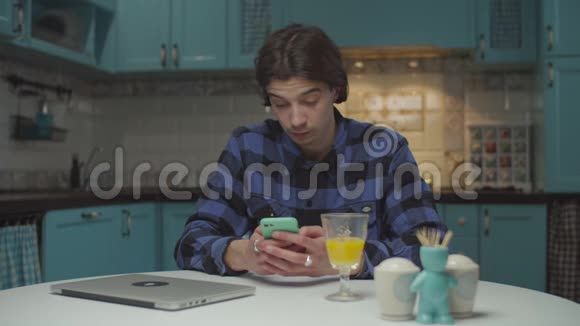 20多岁的年轻人用手机上网微笑着坐在蓝色厨房的餐桌旁男性视频的预览图