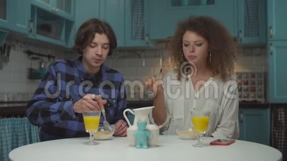 年轻快乐的20多岁夫妇穿着休闲服装在舒适的蓝色厨房里吃早餐男人和女人一起吃玉米片视频的预览图