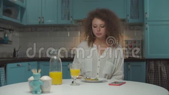 年轻的20多岁卷发女人独自在厨房吃饭女性在家吃晚饭喝橙汁视频的预览图