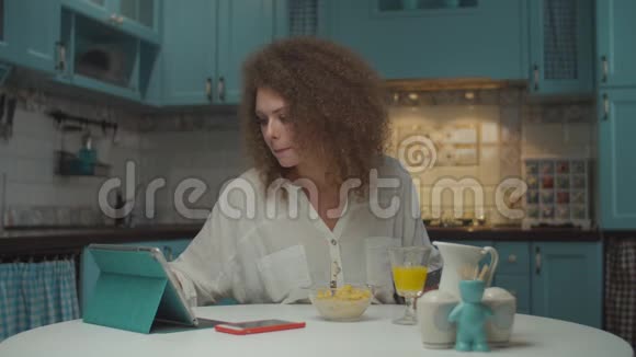 年轻的20多岁卷发女人独自吃早餐在蓝色厨房的平板电脑上在线购物女性视频的预览图