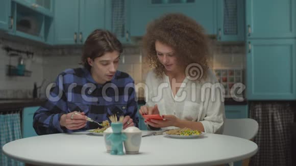 一对20多岁的年轻夫妇在蓝色的厨房里用小玩意儿吃饭男人和女人用手看手机视频的预览图