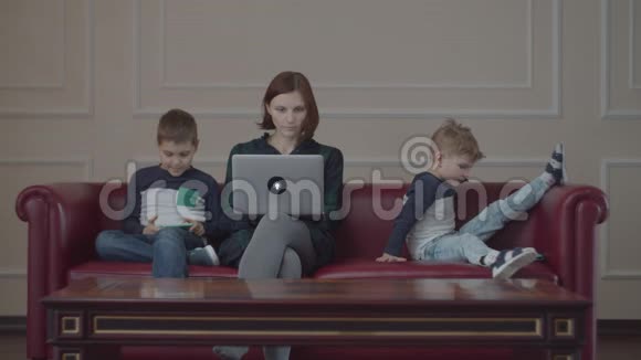 30岁的年轻母亲带着笔记本电脑两个男孩穿着同一件衣服用小工具坐在沙发上家庭在线视频的预览图