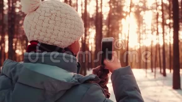 一位年轻漂亮的女士用手机在冬日的雪日落森林拍照视频的预览图