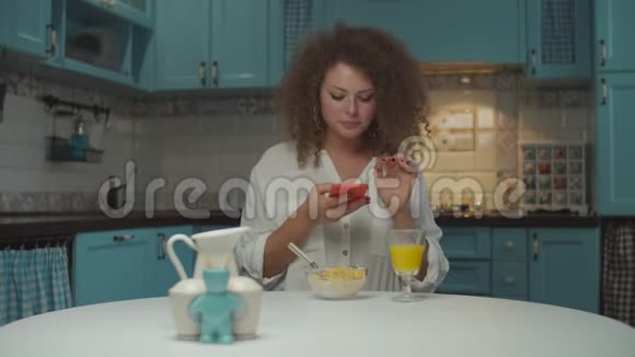 年轻的20多岁卷发女人独自吃早餐手里拿着手机在厨房里女性拍照视频的预览图