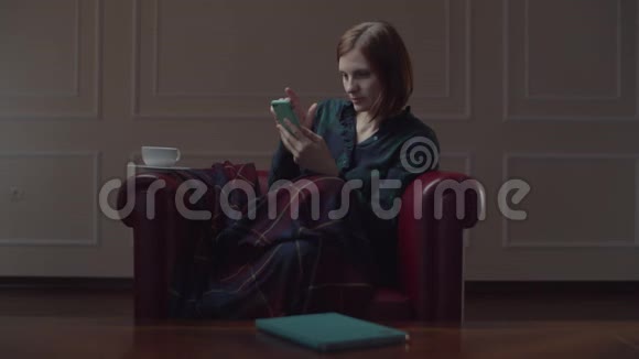 30多岁的年轻女性使用手机和咖啡坐在舒适的扶手椅上有格子呢独自在家工作的女性视频的预览图