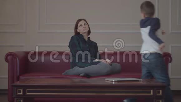 精疲力竭的年轻母亲坐在家里的红色沙发上两个吵闹的孩子在沙发上跑来跑去疲倦的女人头痛视频的预览图