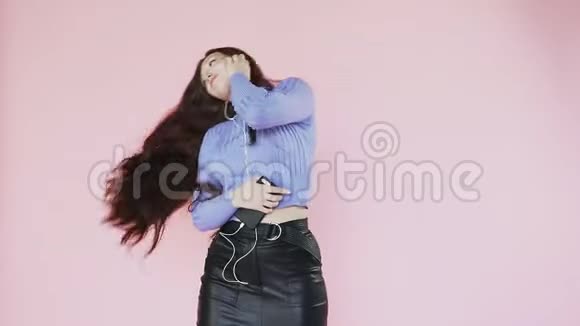 戴着耳机的漂亮红发女孩听音乐和跳舞在粉红色的背景上慢动作视频的预览图