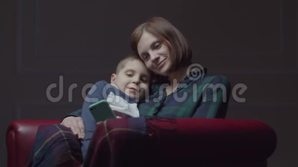 30多岁的年轻母亲带着上学的儿子使用手机坐在舒适的扶手椅上家庭在家里玩小玩意视频的预览图