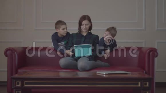 30多岁的母亲坐在沙发上用平板电脑两个穿着同样衣服的男孩和妈妈一起看小玩意视频的预览图