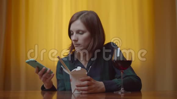年轻的30多岁的女性成年人用筷子吃亚洲盒饭在家里用手机喝葡萄酒一个女人视频的预览图