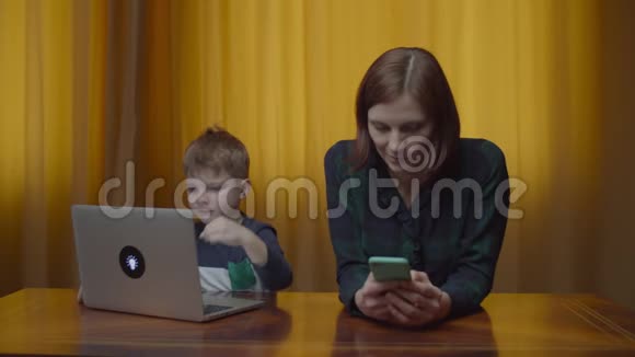 金发学龄前男孩在笔记本电脑上打字母亲带着手机坐在家里的桌子上儿童在线学习视频的预览图