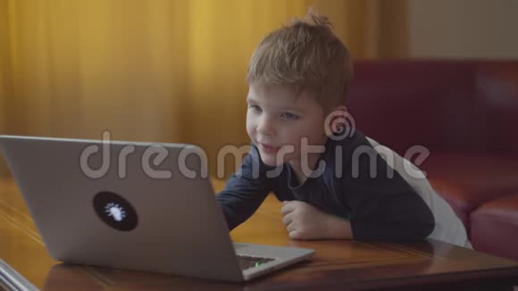 金发的学龄前男孩在笔记本电脑上打字微笑着看着电脑屏幕孩子在家坐在网上学习视频的预览图