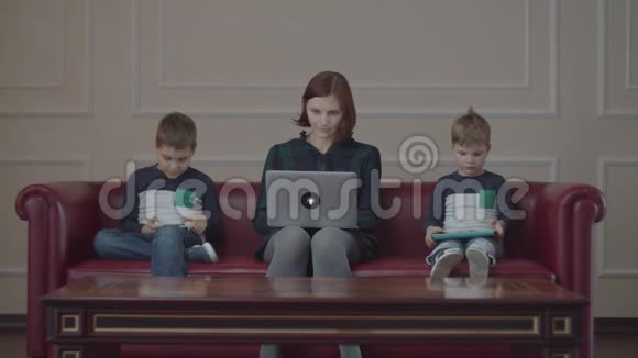 30岁的年轻母亲带着笔记本电脑两个男孩穿着同一件衣服用小工具坐在沙发上家庭在线视频的预览图