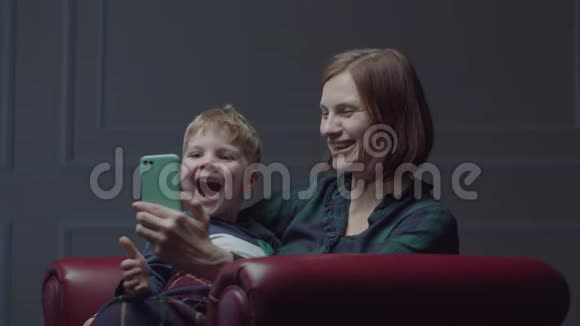 30多岁的年轻母亲带着学龄前儿童使用手机坐在舒适的扶手椅上一家人玩小玩意视频的预览图