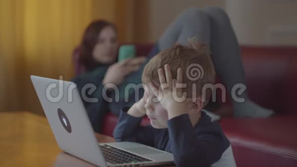 金发的学龄前男孩在笔记本电脑上打字微笑着看着电脑屏幕妈妈拿着手机躺在沙发上视频的预览图