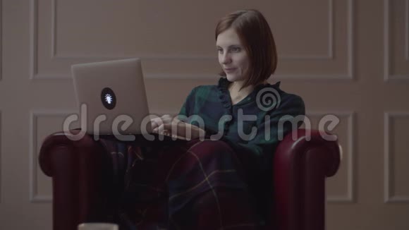 30多岁的年轻女性坐在有格子的舒适扶手椅上用笔记本电脑打字女性独自在家工作视频的预览图
