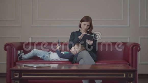 年轻的30多岁的母亲拿着平板电脑男孩用小玩意躺在沙发上家庭在线观看内容视频的预览图