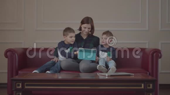 30多岁的母亲坐在沙发上用平板电脑两个穿着同样衣服的男孩和妈妈一起看小玩意视频的预览图