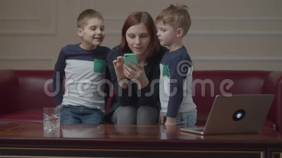 年轻的30岁的母亲坐在沙发上用手机两个儿子和妈妈一起看智能手机家庭在线视频的预览图