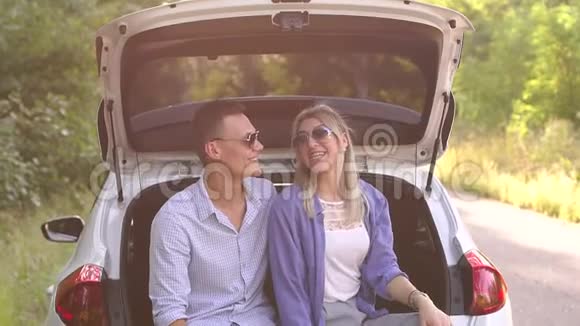 年轻的幸福夫妇戴着太阳镜坐在汽车后备箱里他们正在旅行视频的预览图