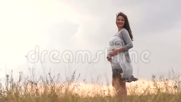 美丽的孕妇长着棕色的头发在日落时站在田野里视频的预览图
