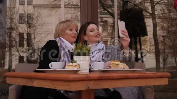 老年妇女和她的成年女儿在人行道咖啡馆的智能手机上自拍视频的预览图