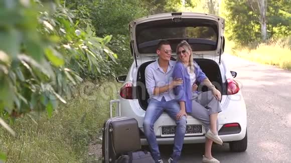 年轻的幸福夫妇戴着太阳镜坐在汽车后备箱里他们正在旅行视频的预览图