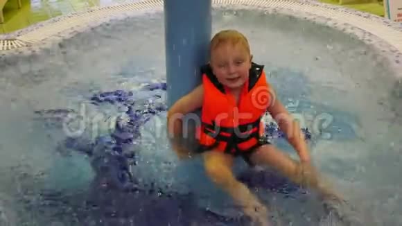 一个穿着橙色救生衣的小孩子坐在水上公园的儿童游泳池里视频的预览图