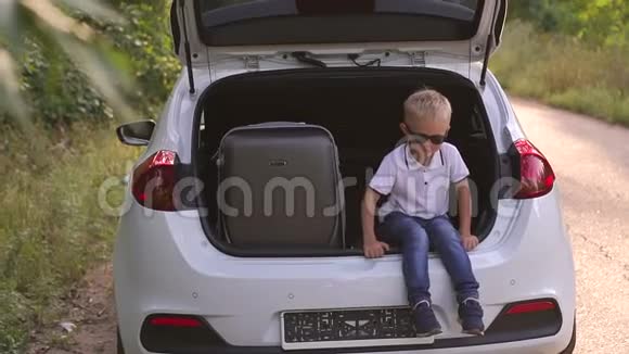 一个戴着太阳镜的小男孩坐在一个灰色手提箱附近的汽车后备箱里视频的预览图