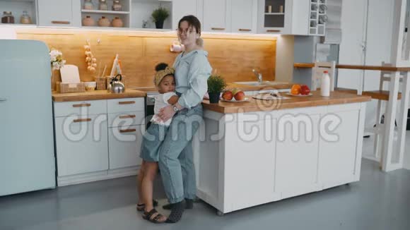 可爱的年轻女性妈妈和非裔美国女孩快乐的女儿抱在明亮的厨房里冰箱视频的预览图