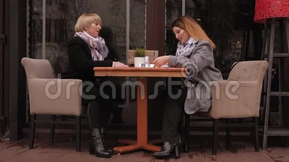 一位年迈的母亲和一位成年的女儿坐在街头咖啡馆的一张桌子旁桌上有菜单视频的预览图