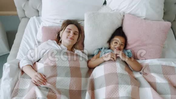 从上到下看美丽的年轻妈妈和快乐的非裔美国女儿躺在床上微笑的母亲视频的预览图