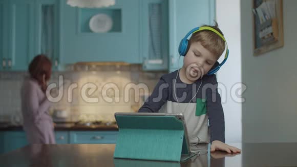 金发的学龄前男孩戴着彩色耳机在厨房的桌子上使用平板电脑母亲使用手机视频的预览图