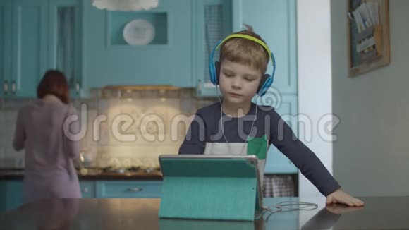 金发的学龄前男孩戴着彩色耳机在厨房的桌子上使用平板电脑妈妈在家里视频的预览图