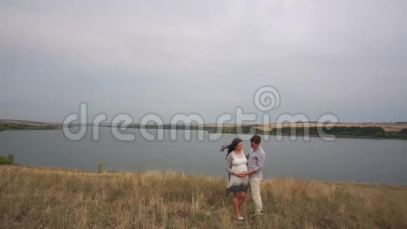 一个男人轻轻地抚摸着站在一个大湖附近的怀孕妻子的肚子视频的预览图