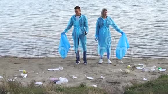 两个穿着蓝色长袍和手套的志愿者站在河岸上的垃圾中视频的预览图