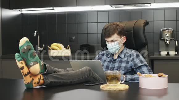 男子使用保护性医疗面罩同时在家里工作在笔记本电脑上以防止冠状病毒感染covid19男性穿着视频的预览图