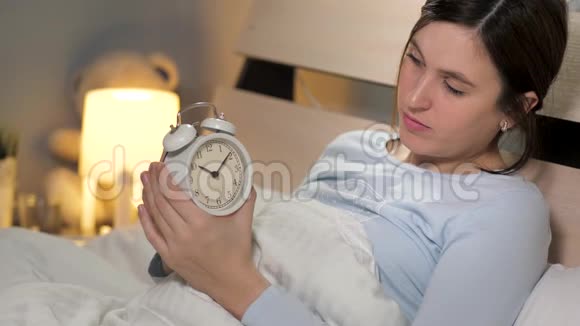 女孩设置闹钟一个年轻漂亮的女人独自躺在卧室里躺在床上手里拿着一个闹钟视频的预览图