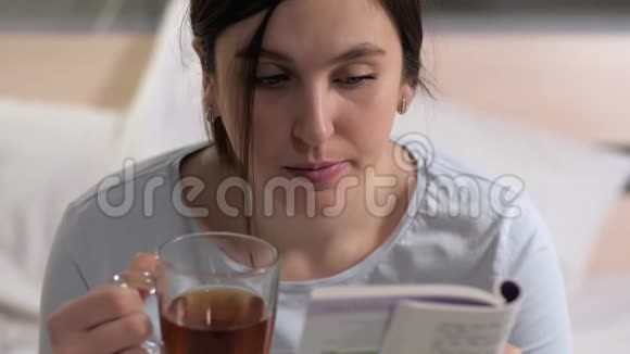 女孩在看书年轻迷人的白种人女人晚上坐在床上看书喝红茶视频的预览图