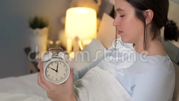 女孩上床睡觉了年轻迷人的白种人女人躺在床上设置老式闹钟放在床头柜上视频的预览图