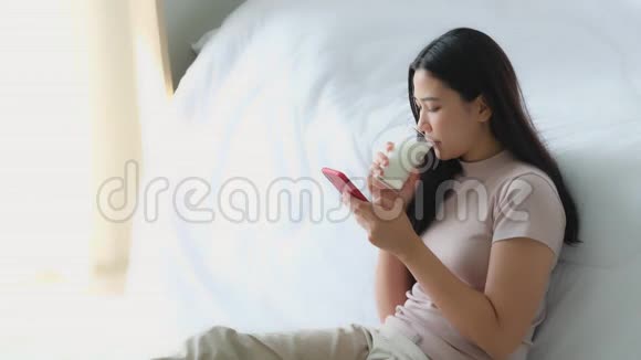 美丽的亚洲年轻女性坐在床边用电话上网购物视频的预览图