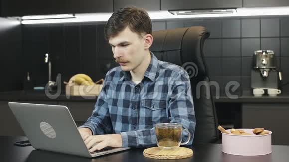 男性自由职业者在家使用笔记本电脑白种人留胡子的男人在厨房的笔记本电脑上做在线工作时喝茶任何人视频的预览图