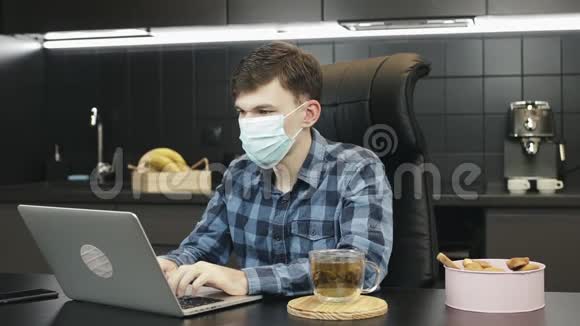 在家中使用笔记本电脑时使用保护性医疗面罩的人穿着医用口罩在厨房的笔记本上打字视频的预览图