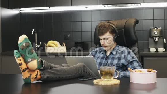 年轻的男性在耳机听音乐和工作笔记本电脑从家里人放松并在家庭办公室工作盖盖视频的预览图