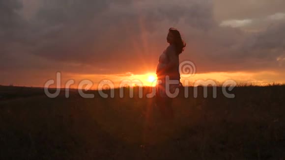 日落时分在一片狂风大作的田野里穿着连衣裙的孕妇剪影视频的预览图