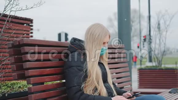 戴着医疗面具的金发女孩坐在长凳上用智能手机视频的预览图
