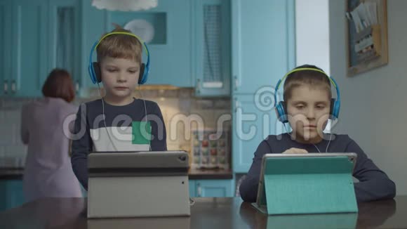 两个兄弟戴着彩色耳机在厨房的桌子上使用平板电脑妈妈有家庭厨房习惯视频的预览图